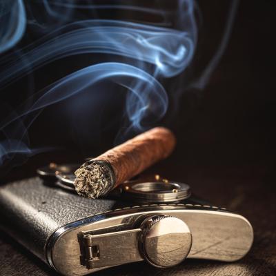 Burning one cigar on leather hip-flask and cutter on wooden table